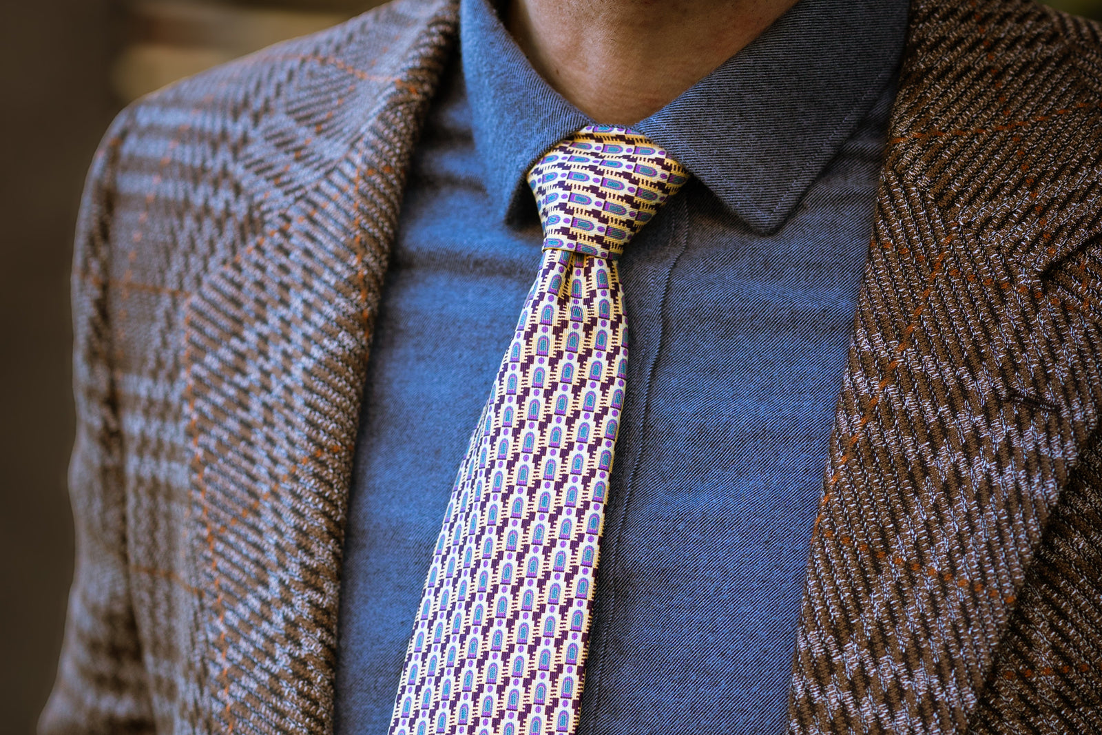 Man wearing a patterned silk tie with a textured brown jacket and blue shirt.